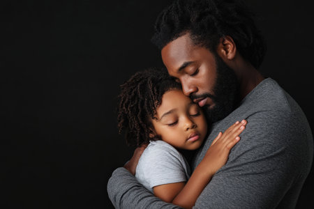 Tender moment between African male adult and child in warm embrace against dark background.の素材