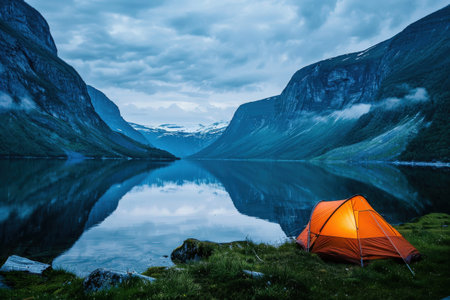 Serene camping by mountain lake at twilight with orange tent reflected in water.
