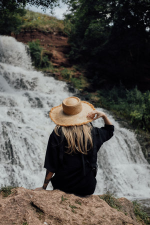 Blonde woman wearing straw hat relaxing by waterfall in nature.の写真素材