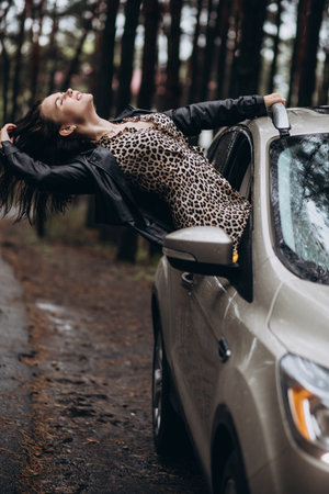 Joyful woman in leopard dress leaning out of car in a forest setting.の写真素材
