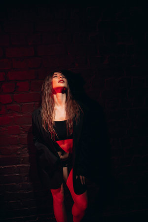 Dramatic red lighting frames woman in dark stylish attire against brick wall.