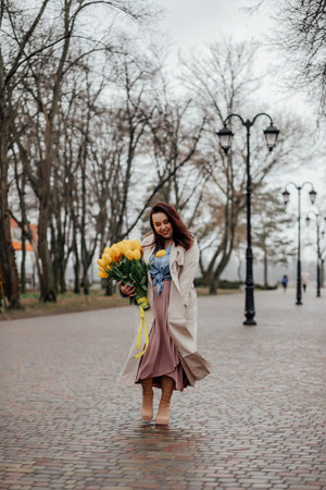 Brunette girl with long hair in beige coat with a bouquet of yellow tulips.の写真素材
