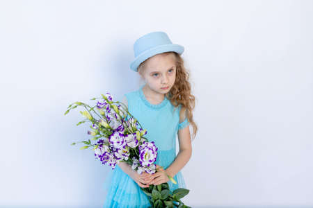 sad girl 5-6 years old in a blue dress and hat on a white isolated background holding a bouquet of flowers, space for textの写真素材