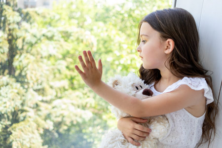 Charming little girl 5-6 years old hugs a Teddy bear and looks out the window