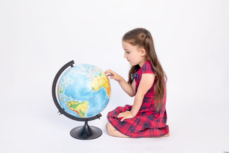 a school girl sits on a white isolated background with a globe and searches for countries with her...