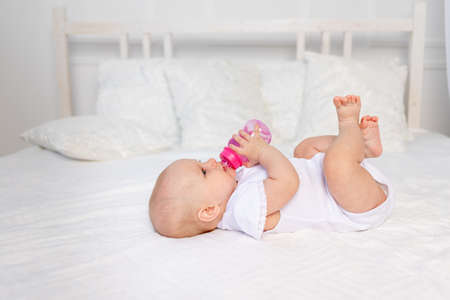 a 6-month-old baby lies on a white bed and drinks milk from a bottle, baby food concept, text spaceの写真素材
