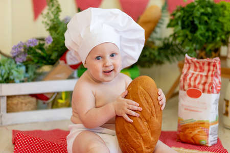 portrait of a baby in a cook's hat with bread in his hands in a beautiful photo zone with flour and vegetables, a cook's child, a child eating bread, preparing foodの写真素材