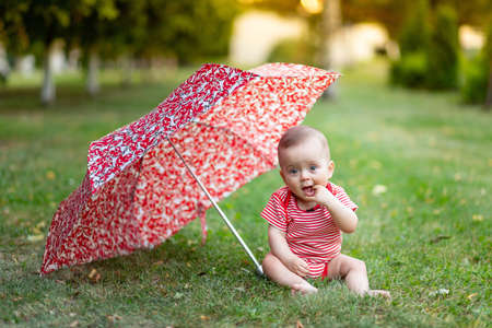 a little girl 8 months old sits on a green lawn in the summer under a red umbrella at sunset.の写真素材