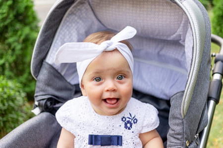 a small child a girl of 7 months sits in a stroller in the summer in a white dress and shows her...