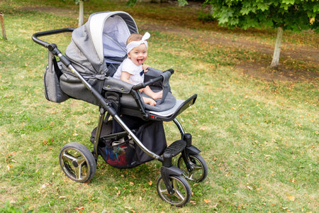 a small child a girl of 7 months sits in a stroller in the summer in a white dress and looks at the...