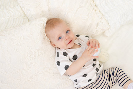 baby 8 months old lying on the bed at home in pajamas with a bottle of milk, baby food concept, baby...