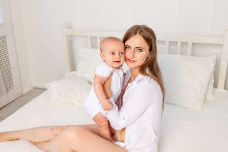 mother holds baby 6 months sitting on a white bed at home and looking at the camera, a place for textの写真素材