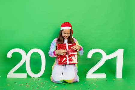 a cute little girl on a green isolated background in a bright suit and Santa hat with a gift in her hands among the numbers 2021. Space for text. New year and Christmas conceptの写真素材