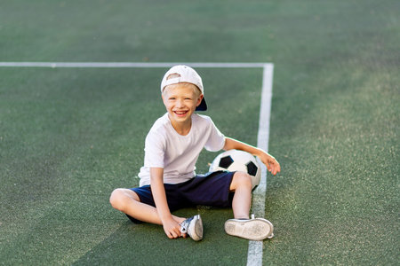 a blond boy in a cap in a sports uniform sits on a football field with a soccer ball, sports...