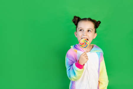 a cute girl is licking a large Lollipop in a bright suit on a green isolated background. Space for text.の写真素材