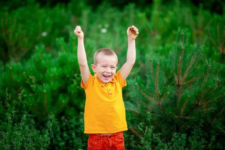 a happy child a boy in orange clothes stands in the green grass in summer and raised his hands upの写真素材