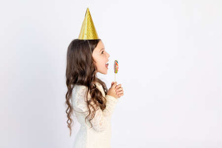 A cute little girl is eating a large Lollipop in a festive hat on a white isolated background. Space for text. Little girl celebrates birthday, holiday conceptの写真素材