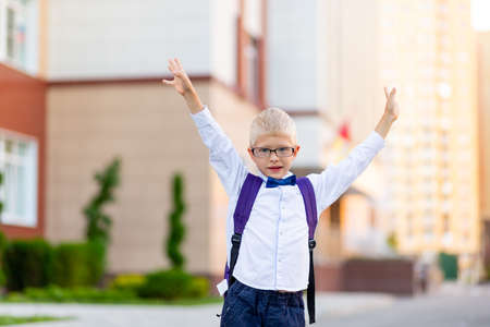 a happy schoolboy boy with blond glasses and a backpack is standing at the school and is happy with his hands up. Day of knowledgeの写真素材