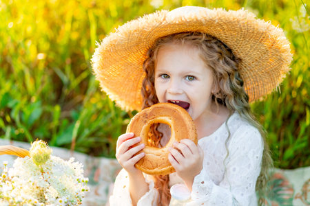 beautiful little girl sitting in a straw hat in a field and eating a bagel, picnic in a field