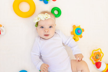 portrait of a small baby girl 6 months old lying on her back on a white bed at home among toys, top...