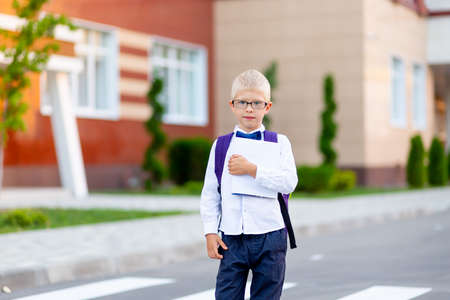 a schoolboy boy with blond glasses with a backpack and a white book is standing at the school. Space for text. Day of knowledgeの写真素材
