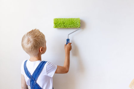 child boy Builder paints white walls with a roller in an apartment, a child makes house repairs,...