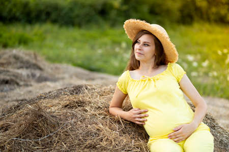 a pregnant woman in dark clothes and a hat sits in a field on straw in summerの写真素材