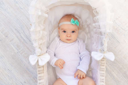 a small baby girl 6 months old in a white bodysuit lies in a beautiful baby bed cradleの写真素材