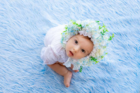 a beautiful little girl sits on a blue fluffy rug in a hat made of flowers and looks at the cameraの写真素材