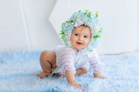 a beautiful little girl is sitting on a blue fluffy rug in a bright room wearing a hat made of flowers and smilingの写真素材