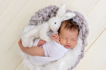 a newborn baby girl is sleeping sweetly in a basket with a soft toy hare in her armsの写真素材