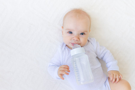 portrait of a small baby girl 6 months old in a white bodysuit lying on her back on a bed with a...