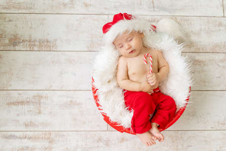 a sleeping child in a Santa costume lies with a candy in his hands on a light wooden background, waiting for the new year and Christmas, space for textの写真素材