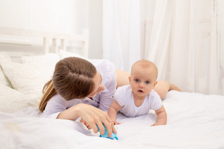 mom playing with baby 6 months lying on a white bed, leisure mom with baby, place for text