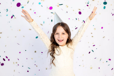 A cute little girl with a cap on a white isolated background with a streamer is happy with her arms...