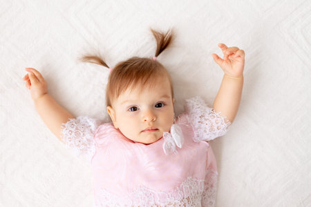 portrait of a little baby girl six months old on a white bed in pink clothes with her hands up
