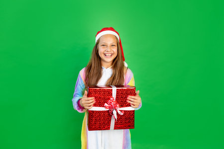 cute little girl on a green isolated background in a bright suit and Santa hat with a gift in her...