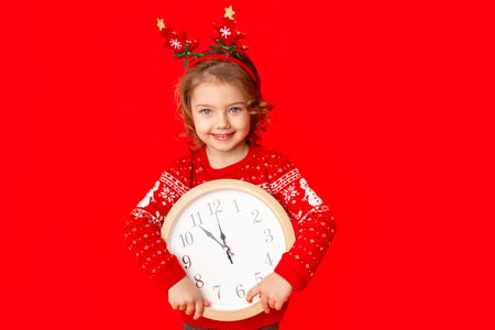 a little girl in winter clothes holds a watch on a red background. New year's concept, space for...