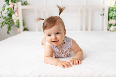 funny baby girl six months old lies in a bright beautiful room on a white bed in a lace bodysuit and...