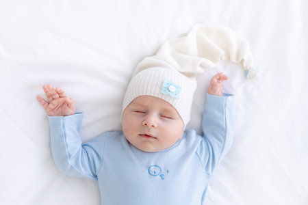 baby boy sleeps on the bed lying on his back in blue pajamas with his hands up, healthy newborn...