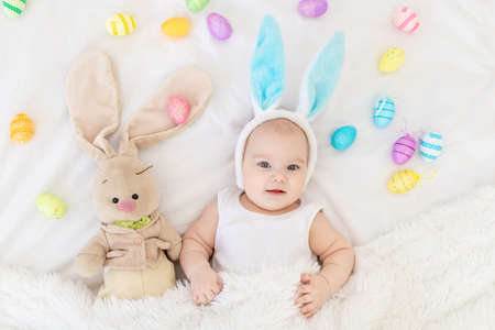 A baby boy with rabbit ears on his head is lying in a crib with a bunny toy and Easter eggs, a cute funny smiling little baby. Easter conceptの写真素材