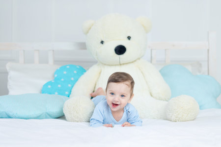 happy smiling baby boy on the bed with a big teddy bear in a blue bodysuit, cute little baby in the bedroomの写真素材
