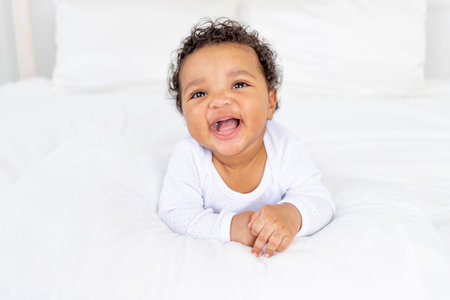 smiling African-American little baby on white bed in bedroom laughing
