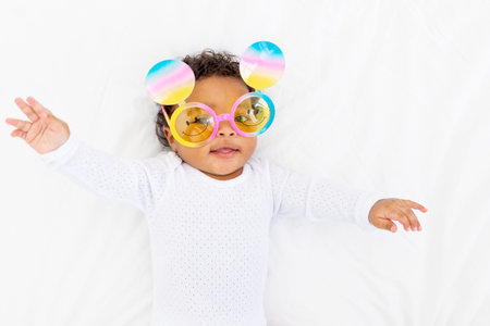 An African-American little child in funny glasses lies on a white bed and smiles