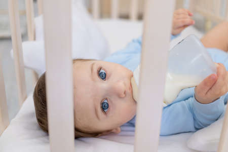 baby boy eating from milk bottle in crib in blue bodysuit, cute little baby in bedroom, baby food conceptの写真素材