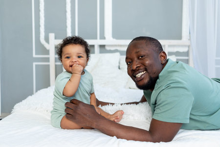 happy smiling African american dad with baby son on bed at home cuddling and playing, happy family,...