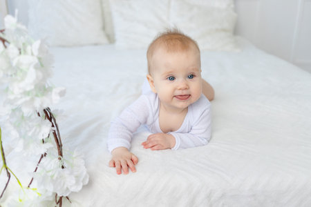 baby girl six months old shows tongue lying on white bed in bedroomの写真素材