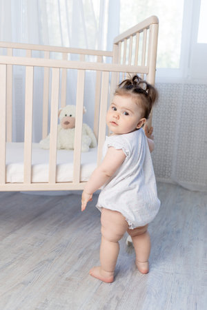 cute baby girl stands at the side of the crib holding on to it with her hands in a white bodysuit,...