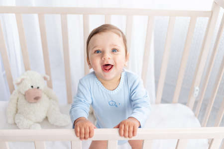 happy baby boy stands in the crib in the nursery and smiles or laughsの写真素材