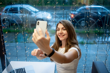 a smiling freelancer girl takes a selfie on the phone or makes a video call, a working day of a free...